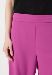 Fuchsia wijde broek met een zachte textuur en een hoge tailleband, met minimale naden voor een cleane uitstraling. Hand rustend op de stof.