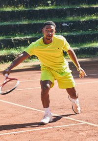 Jugador de tenis con una camiseta y pantalones cortos amarillos brillantes, concentrado en un golpe de derecha, con zapatillas de tenis blancas en una cancha de tierra batida roja.