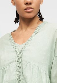 Jeune personne aux cheveux foncés tressés portant un chemisier vert clair texturé avec un bord en dentelle florale le long du col en V et de la couture avant.