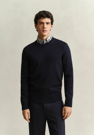 Jeune homme aux cheveux bouclés et foncés portant un pull noir par-dessus une chemise à carreaux, debout devant un fond clair uni.