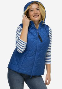 Prošívaná modrá vesta s kapucí a žlutou podšívkou, má dvě přední kapsy na zip a je spárována se pruhovanou košilí s dlouhým rukávem.