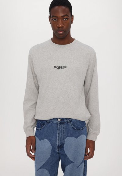 Gray long-sleeve shirt with "SELF LOVE CLUB OPEN 24/7" embroidered in black; worn with blue jeans featuring light blue heart-shaped patches.