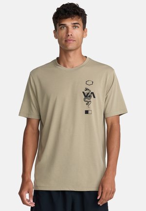 Giovane uomo con capelli corti e ricci che indossa una maglietta beige con un serpente nero e simboli geometrici sul petto, in piedi davanti a uno sfondo bianco.