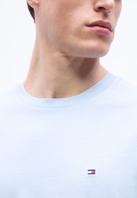 Κοντινό πλάνο του κάτω μέρους του προσώπου και του λαιμού ενός ατόμου που φορά ένα ανοιχτό μπλε μπλουζάκι με ένα μικρό κόκκινο, λευκό και μπλε λογότυπο στο στήθος.