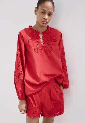 Kvinde iført en rød langærmet top og matchende shorts med blomstrede blondeudskæringsdetaljer, stående ved en hvid væg.