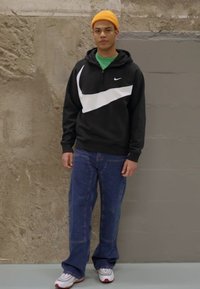 Čierno-biela mikina Nike s dizajnom na štvrť zips, voľné modré džínsy a biele tenisky s červenými akcentmi. Oranžová pletená čiapka.