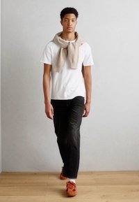 White cotton t-shirt, black jeans, beige sweater draped around neck, and orange sneakers. Wood floor and gray wall background.