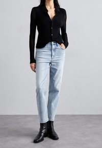 Žena v černé košili s dlouhým rukávem a knoflíky, světlých modrých džínech s vysokým pasem a černých kotníkových botách, stojící s jednou rukou v kapse.