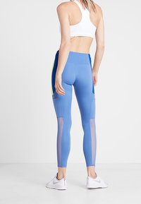 Žena v modrých sportovních legínách s mřížkovanými panely a bílou sportovní podprsenkou, stojící zády k fotoaparátu na čistém bílém pozadí.