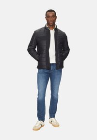 Chaqueta acolchada negra con un cuello alto, que presenta un panel oscuro y un logo. Combinada con jeans azules y zapatillas beige con detalles verdes.