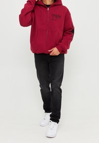Persona con una sudadera burgundy con cremallera, texto negro y un gráfico de escorpión en la manga, jeans negros y zapatillas blancas.