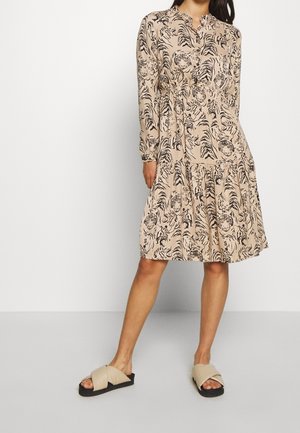 Femme portant une robe beige mi-longue avec un imprimé tigre noir et des manches longues, associée à des sandales slip-on beiges avec semelles noires.