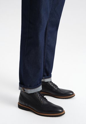Stivali neri in pelle con lacci indossati con jeans di denim blu scuro risvoltati alle caviglie, mostrati su uno sfondo bianco semplice.