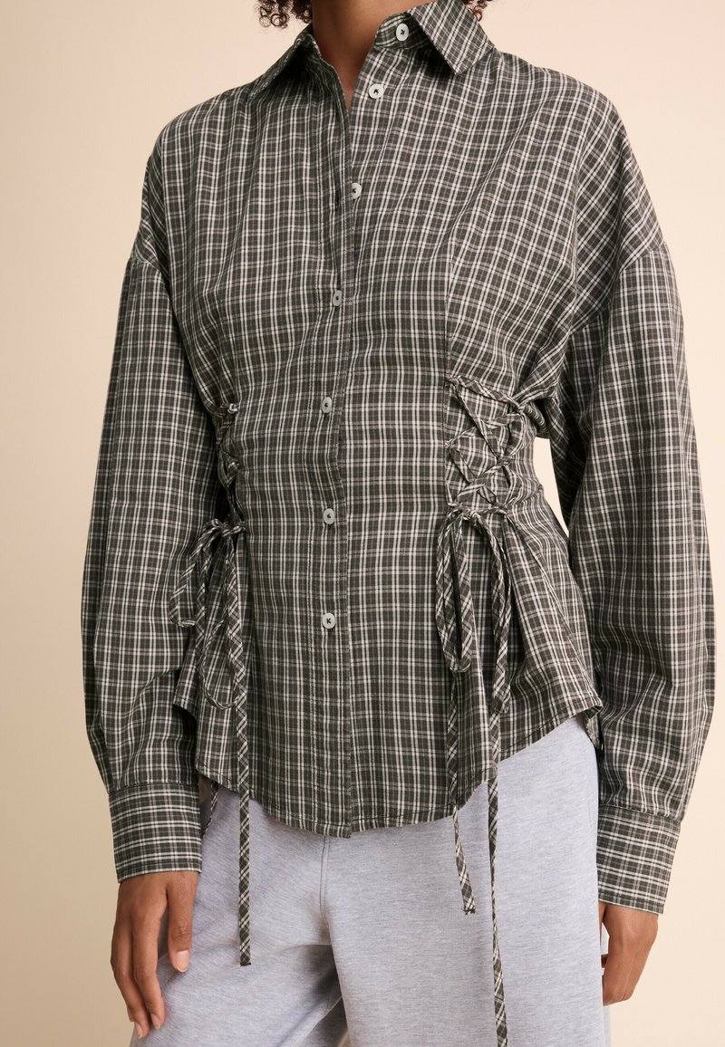 Femme portant une chemise à carreaux verte et blanche boutonnée avec des détails lacés sur les côtés et des manches longues, associée à un pantalon gris clair.