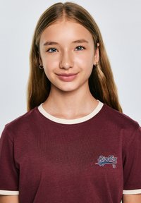 Burgundijas t-krekls ar krēmveida apaļu izgriezumu, ar zilu logo kreisajā krūtī. Gluda auduma tekstūra un īsās piedurknes.