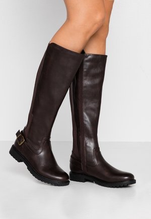 Femme portant des bottes hautes en cuir marron foncé avec des bouts arrondis, des boucles près du talon et des semelles noires robustes, sur un fond clair uni.
