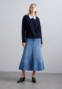 Donna che indossa un cardigan blu navy con bottoni e colletto bianco, una gonna in denim a trapezio lunga e mule nere con tacco, in piedi contro un muro chiaro e uniforme.