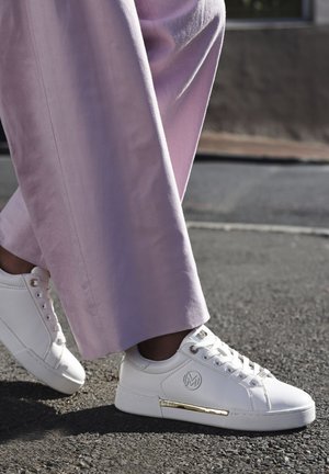 Witte leren sneakers met een gouden accentstreep, ronde neus en vetersluiting, gecombineerd met brede lavendelpantalons in een straatsetting.