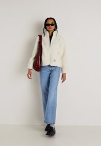 Giacca in pile bianca con due tasche frontali, abbinata a jeans a gamba larga di un blu chiaro. Occhiali da sole scuri e una borsa a tracolla bordeaux completano l'outfit.