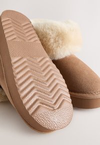 Pantofole in pelle scamosciata marrone con una morbida fodera in pelliccia color crema, suole di gomma testurizzate con motivo a zigzag e punta liscia e arrotondata.