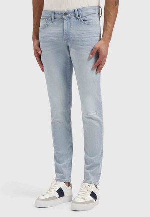 Onderlichaam van een persoon die lichtblauwe slim-fit jeans en witte sneakers met zwarte zijdestrepen draagt, staand tegen een effen achtergrond.