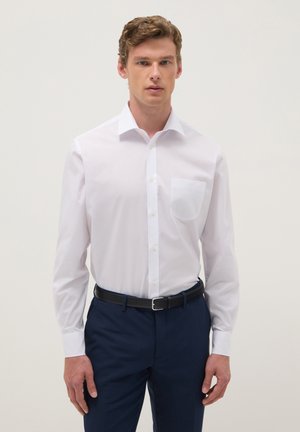 Jeune homme portant une chemise blanche à boutons avec une poche poitrine, un pantalon bleu foncé et une ceinture noire, debout devant un fond clair.