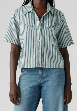 Personne portant une chemise à manches courtes rayée verticalement bleu et blanc avec un jean en denim bleu.