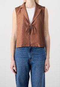 Brun ärmlös väst med knyt framtill, snibbhals och texturerad tyg, kombinerad med högmidjade blå denimjeans.
