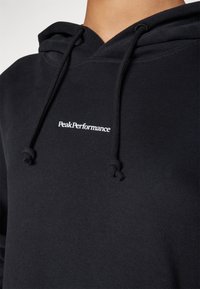 Svart hoodie av mjukt tyg, med en känguruficka, dragsnören i huvan och en liten vit "PeakPerformance"-logga på bröstet.