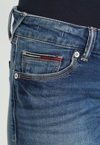 Tmavě modré džíny z denimu s vybledlým vzhledem, které mají malou přední kapsu s červenými a modrými pruhovanými detaily a stříbrným knoflíkem.