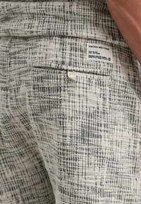 Hellgraue, strukturierte Hose mit einem schwarzen Karomuster, ausgestattet mit einer Gesäßtasche mit einem weißen Knopf und einem Stofflabel in der Nähe der Taille.