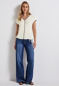 Ljus krämfärgad blus med svart piping, korta ärmar och knytning vid midjan, parat med vida blå denimjeans.