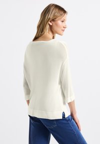 Maglione leggero di colore crema con maniche a tre quarti, texture sottile, spacchi laterali all'orlo, abbinato a jeans blu scuro.