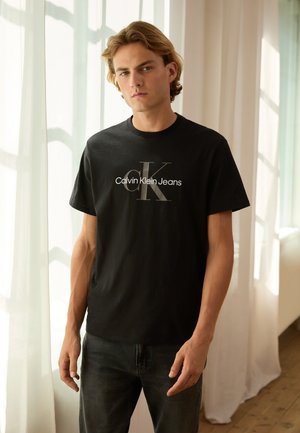 Camiseta de algodón negra con un logo blanco de "Calvin Klein Jeans" y un gráfico estilizado de "CK" en el frente, escote redondo, mangas cortas.