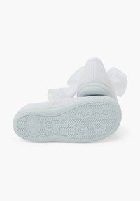 Zapato de tela suave texturizada blanca con punta redondeada, que presenta un gran lazo blanco y una suela de goma con un patrón de olas.