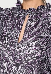 Bluzka we wzory z falbanianym kołnierzem, wycięciem typu keyhole oraz mieszanką czarno-biało-fioletowych falistych wzorów na gładkiej tkaninie.