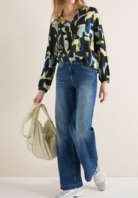 Femme portant un jean bleu à jambes larges, un chemisier imprimé abstrait et coloré, des baskets argentées, tenant un sac à main beige lâche à ses côtés.