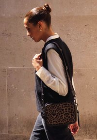 Kabelka se zvířecím potiskem leopardu s černým koženým popruhem. Nošena přes rameno, vedle tmavého vesty přes bílou košili, na neutrálním pozadí.