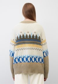 Personne avec de longs cheveux châtain clair portant un pull à motifs crème, marron, jaune et bleu, se tenant devant un fond blanc uni.