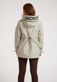 Lichtgrijze waterproof jas met een getailleerde taille, verstelbare capuchon en zwarte "JACKIT" tekst op de kraag. Glad textuur, lange mouwen.