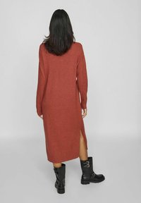 Robe longue tricotée de couleur rouille, avec manches longues et fente latérale, associée à des bottines noires ornées de détails à boucles.