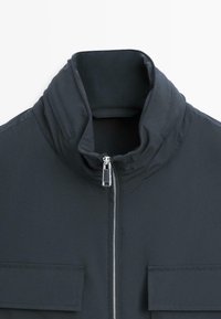 Veste noire avec un col montant et une fermeture éclair argentée à l'avant, dotée de deux poches à rabat horizontales sur la poitrine.