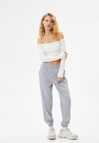 Top blanco de manga larga con los hombros al aire, combinado con joggers gris claro que cuentan con una cinturilla elástica y tobillos ajustados, complementado con zapatillas beigas.