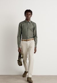 Dunkelgrünes, tailliertes Hemd. Cremefarbene Hosen, brauner Gürtel und olivgrüne Tasche. Braune Schuhe runden das Outfit ab.