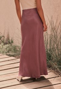 Lang lavendelfarvet nederdel i satin med en glat tekstur, flydende design og en let glans. Båret med lyse sko på en trægulv.