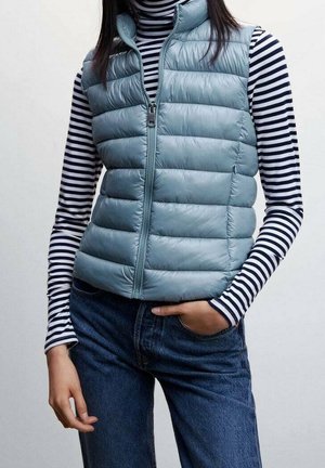 Kobieta w jasnoniebieskiej pikowanej bezrękawniku na czarno-białym pasiastym golfie z długim rękawem, w połączeniu z niebieskimi dżinsami, z ręką w kieszeni.