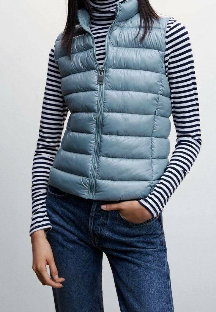 Femme portant un gilet matelassé sans manches bleu clair sur un col roulé à manches longues rayé noir et blanc, associé à un jean bleu, main dans la poche.