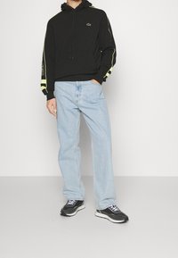 Svart sweatshirt med neongula accenter, avslappnade ljusblå jeans och svarta sneakers med vita snören och en strukturerad design.