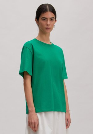 Frau mit zurückgebundenem dunklem Haar, die ein grünes kurzärmeliges T-Shirt und einen weißen Rock trägt, steht vor einem schlichten hellgrauen Hintergrund.