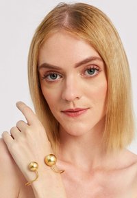 Zlatý kovový náramok s dvoma guľatými akcentmi, hladkým povrchom a otvoreným zakriveným dizajnom, nasadený na zápästí modelu.